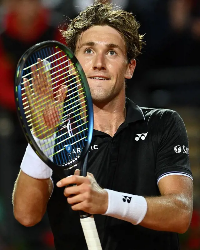 Casper Ruud taps on the net of his racket while looking above.